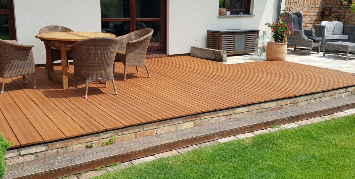 Bambusterrasse mit warmtonigen Dielen vor einem Wohnhaus, darauf stehen vier Korbstühle, im Vordergrund grenzt die Terrasse an einer Rasenfläche. 
